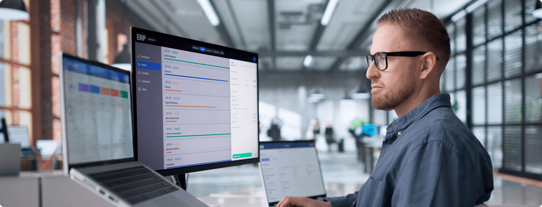 Man with glasses reviewing an ERP system dashboard across multiple monitors and a laptop in a modern open-plan office