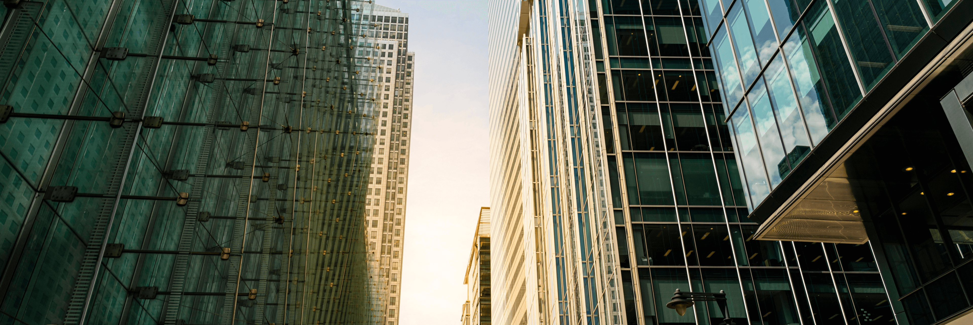 Low-angle view looking up between tall glass and steel corporate skyscrapers toward a warm sky at dusk