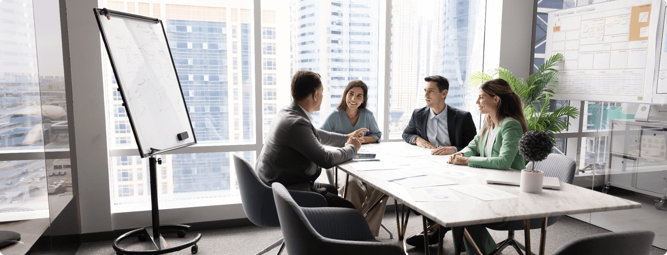 Four business professionals having a discussion around a conference table in a bright high-rise boardroom with a whiteboard and city skyline views