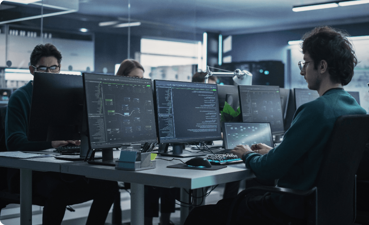 Software development team working at desks with multiple monitors displaying code and system dashboards in a modern low-lit office environment