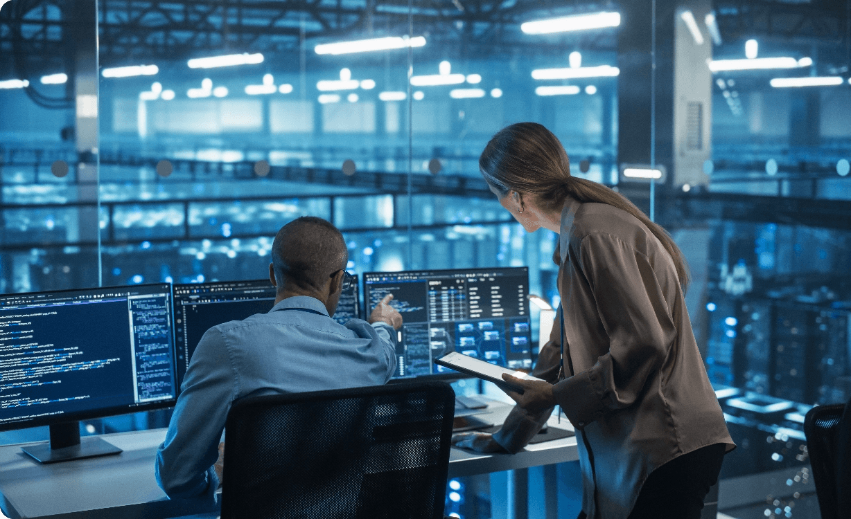 Two IT professionals collaborating at multiple monitors displaying code and data dashboards in a large blue-lit data center operations room