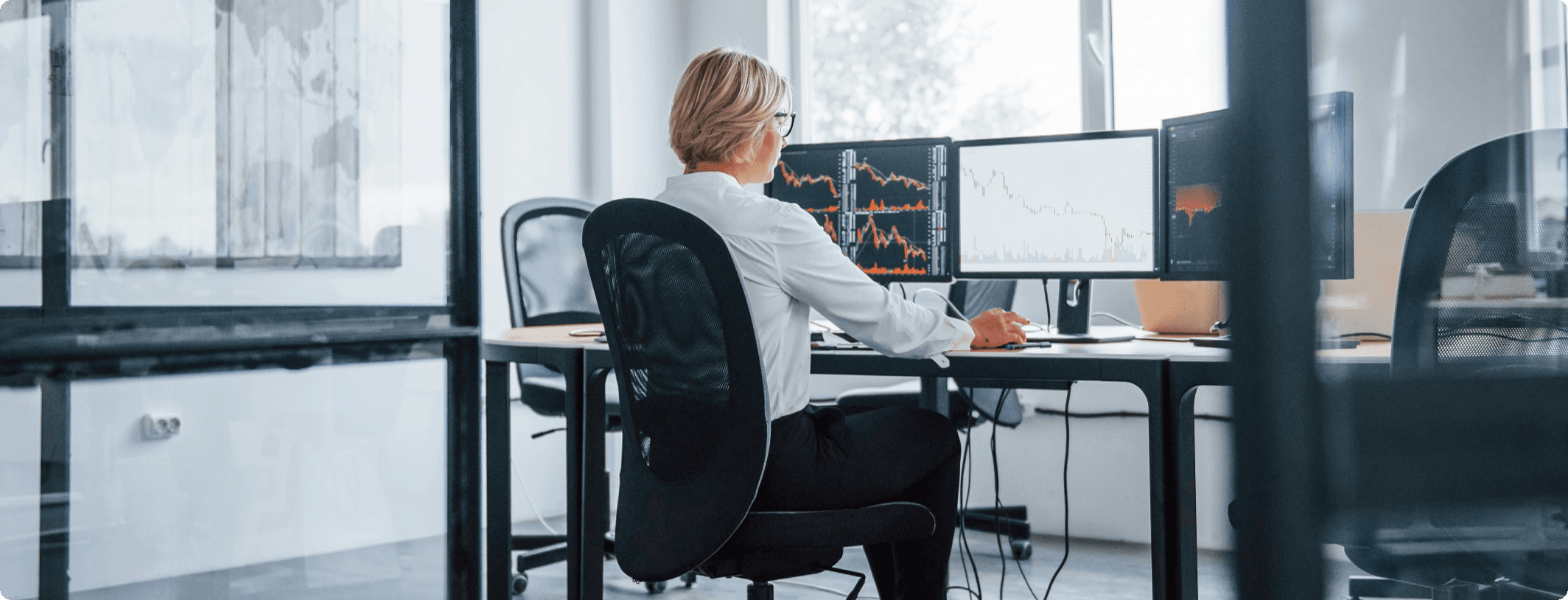 Female financial analyst reviewing declining stock market charts on dual monitors in a bright modern office with large windows