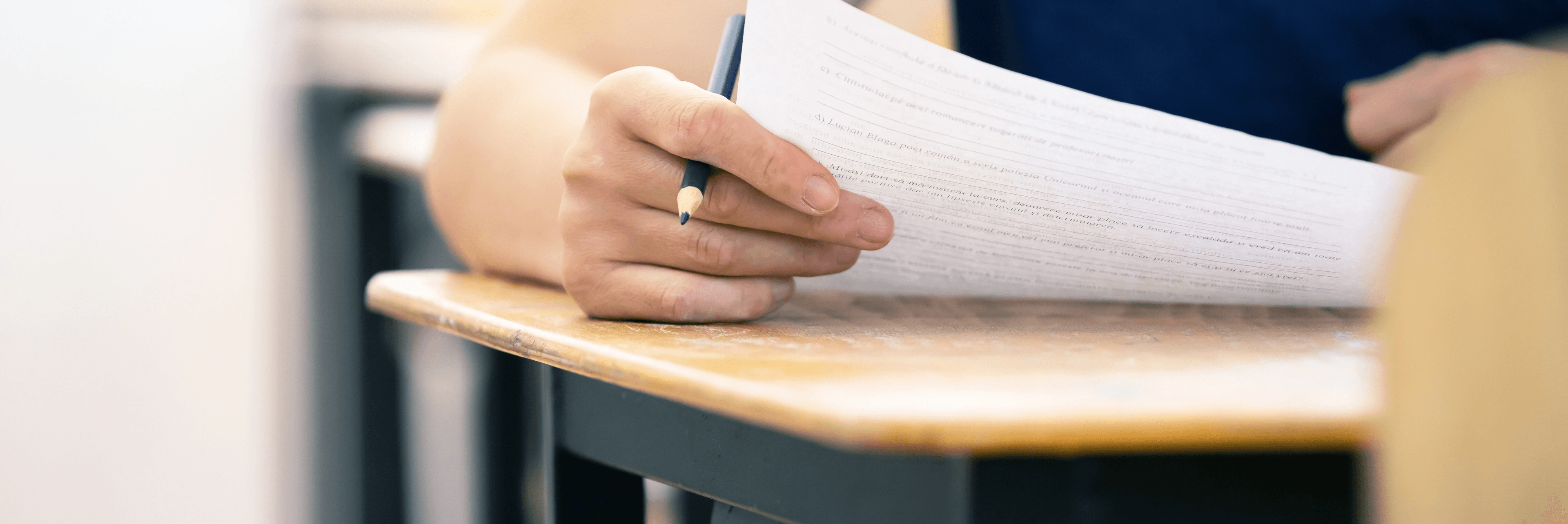 Close-up of a student's hand holding a pencil over a written exam paper on a school desk