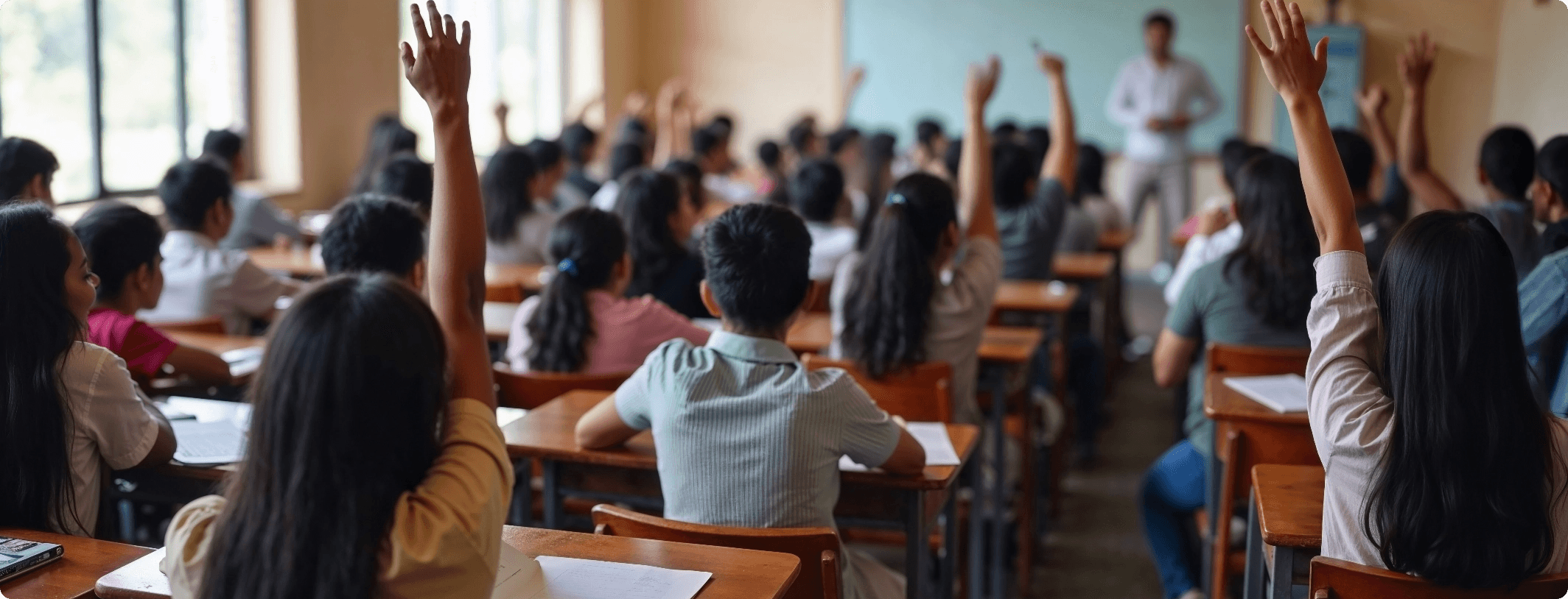 Large classroom of students raising their hands toward a teacher at the front of the room