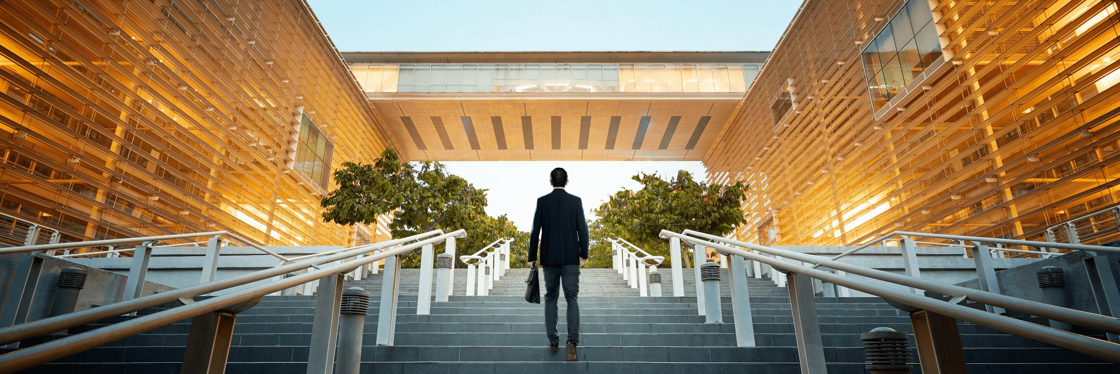 Businessman carrying a bag walking up wide steps toward the entrance of a modern corporate building with warm golden facades at dusk