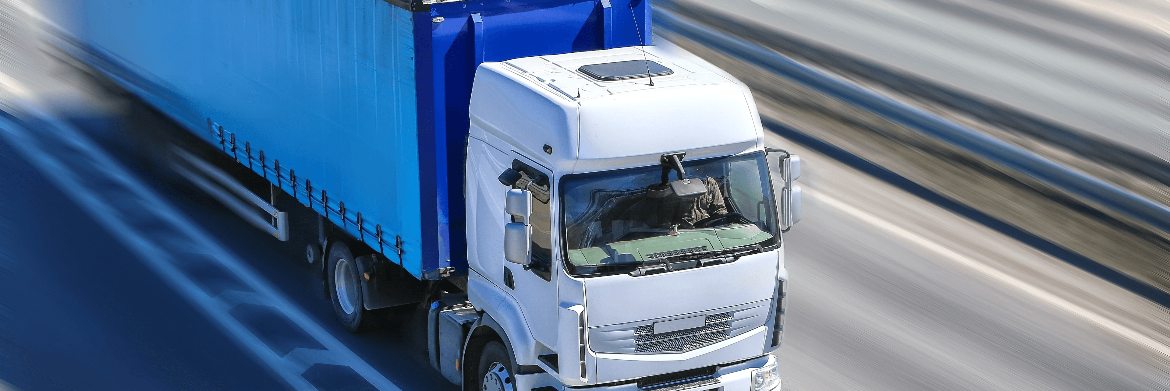 Aerial view of a white semi-truck with a blue cargo trailer traveling at speed on a highway with motion blur on the road surface