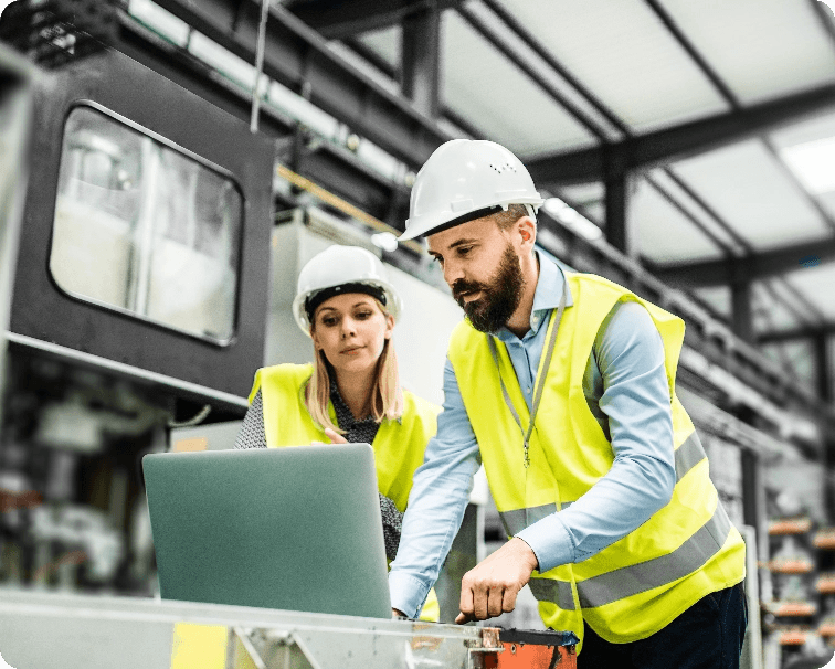 two people in high-vis vests and hard hats using a laptop in an industrial setting